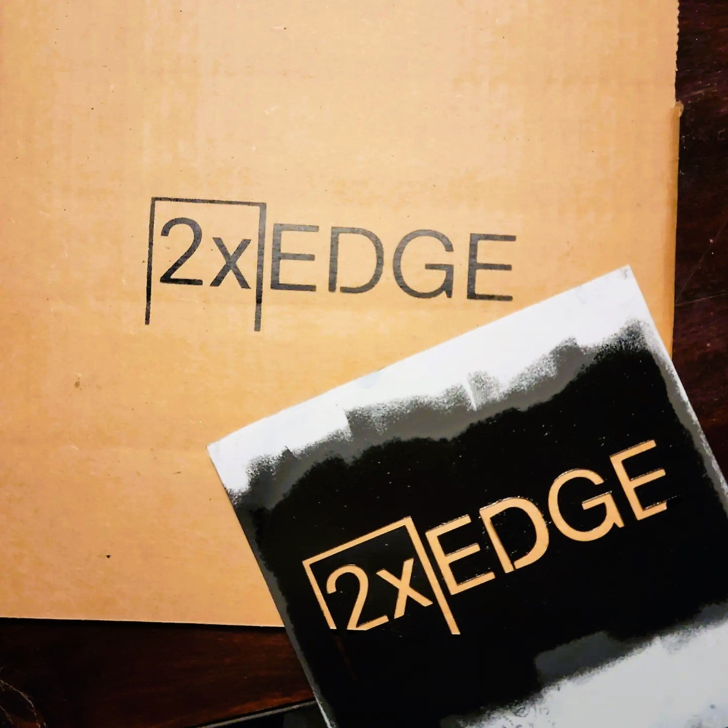 In the background is a cardboard box that has the 2xEDGE logo stenciled across it. In the foreground is the stencil still wet with ink, that was used to stencil the box.