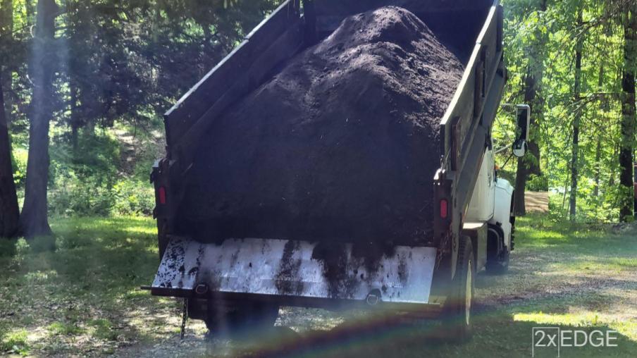 A dump truck unloading mulch onto a driveway with a rainbow in the forefront.