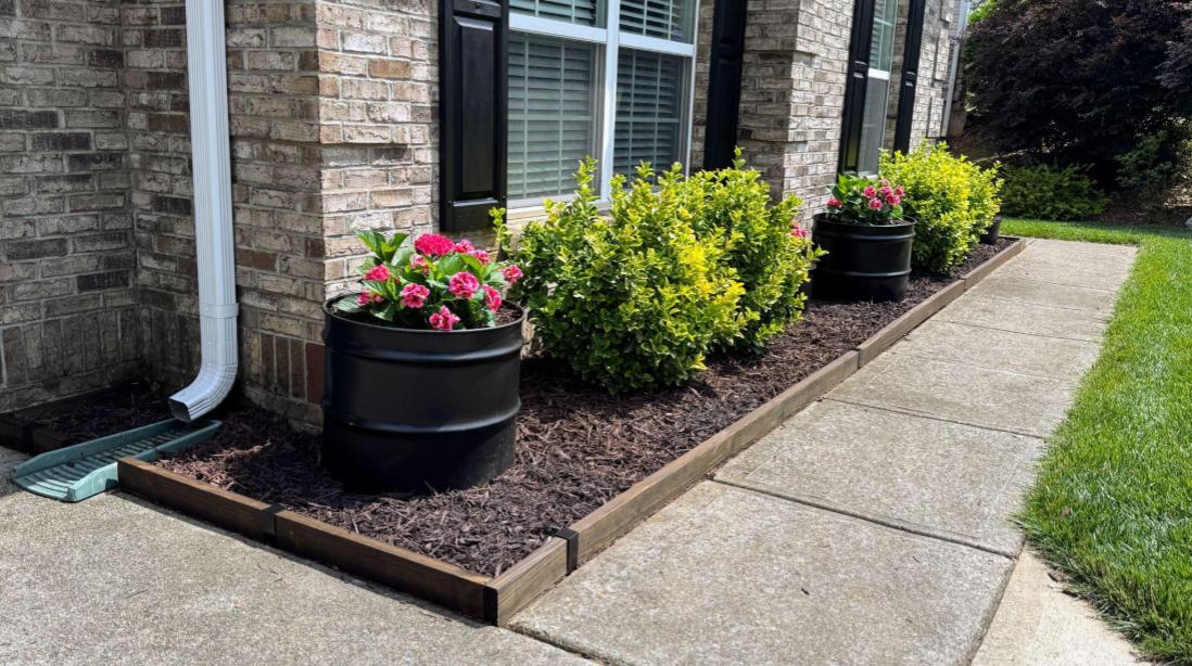 Stained 2x4 flower bed edging installed alongside a concrete sidewalk with dark mulch and black barrel planters.