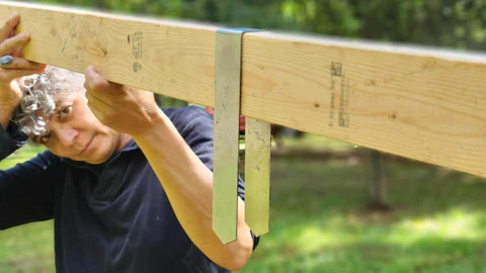 2xEDGE Staples creator Lisa Brooks holding a 2x4 over her head. In the foreground, the 2x4 has an original Rustic Steel 2xEDGE Staple hanging from it to illustrate how the staples wrap around lumber.
