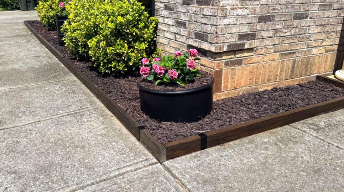 Flower bed corner with stained 2x4 wood edging and satin black 2xEDGE Staples installed alongside concrete sidewalk.