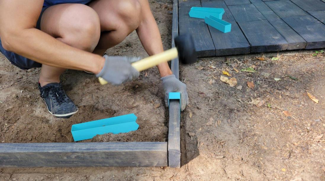 A person using a 16 ounce rubber mallet to install a Robins Egg Blue 2xEDGE Staple over 2x4 lumber stained a dark walnut to be used as garden edging.