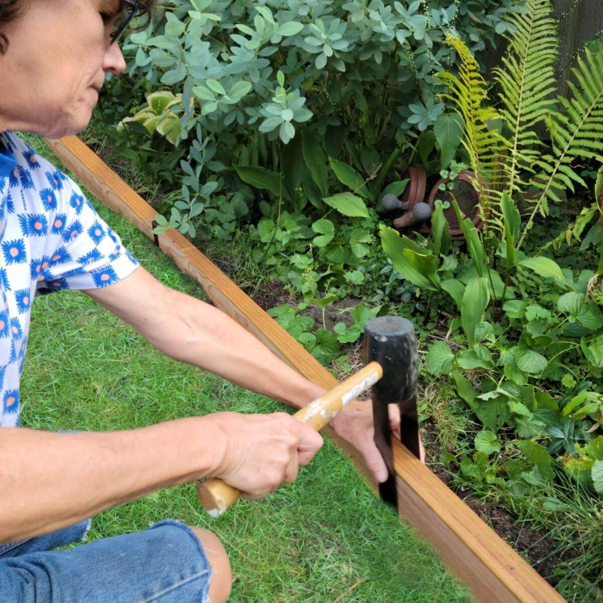 A person using a rubber mallet to install a Satin Black 2xEDGE Staple on a 2x4 to create garden edging between a lawn and a flower bed.