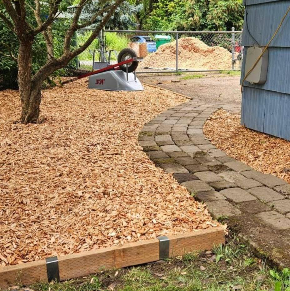 2x4 lumber garden bed edging secured with Rustic Steel 2xEDGE Staples containing a large wood chip mulch bed 
alongside a curved paver walkway with a wheelbarrow and pile of wood chips visible in the background.