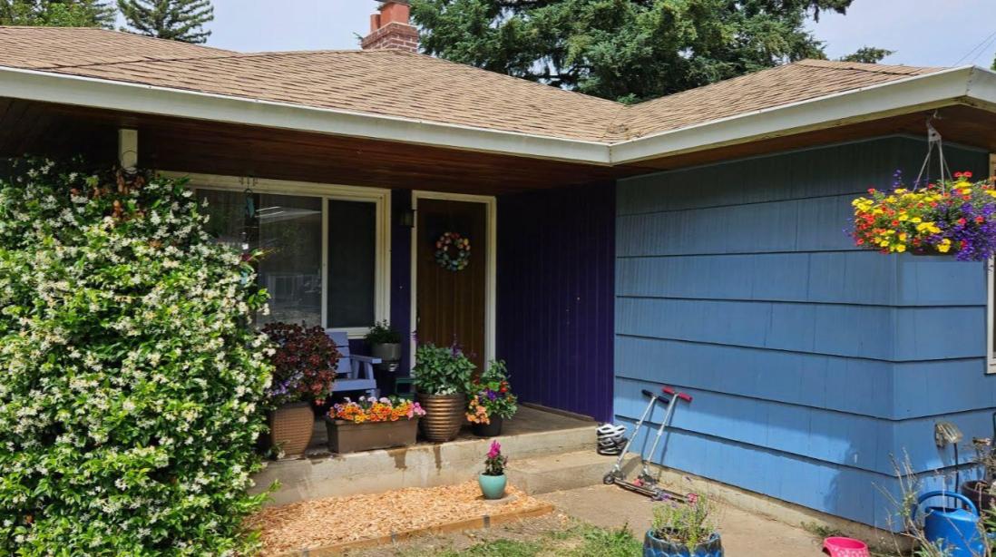 2x4 lumber garden edging alongside a wood chip walkway leading to the front porch of a blue ranch-style home, with colorful container plants and hanging flower baskets.